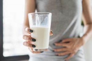 Žena drží pohár mlieka, s bolesťami brucha, ktoré sú jedným z príznakov nedostatku enzýmu laktáza