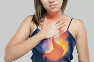 Žena si drží hruď, na hrudi je graficky znázornený plameň a žalúdok, čo symbolizuje pálenie záhy a reflux.