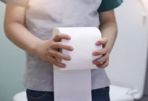 Dieťa drží v rukách rolku toaletného papiera v kúpeľni, symbolizujúc tému stolice ako ukazovateľa zdravia.