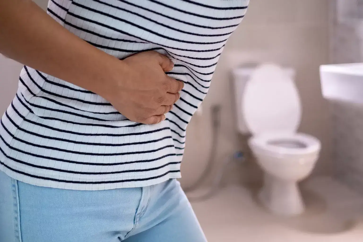 Osoba v pruhovanom tričku si drží brucho, v pozadí je toaleta – znázornenie bolesti brucha a tráviacich ťažkostí typických pri hnačke.